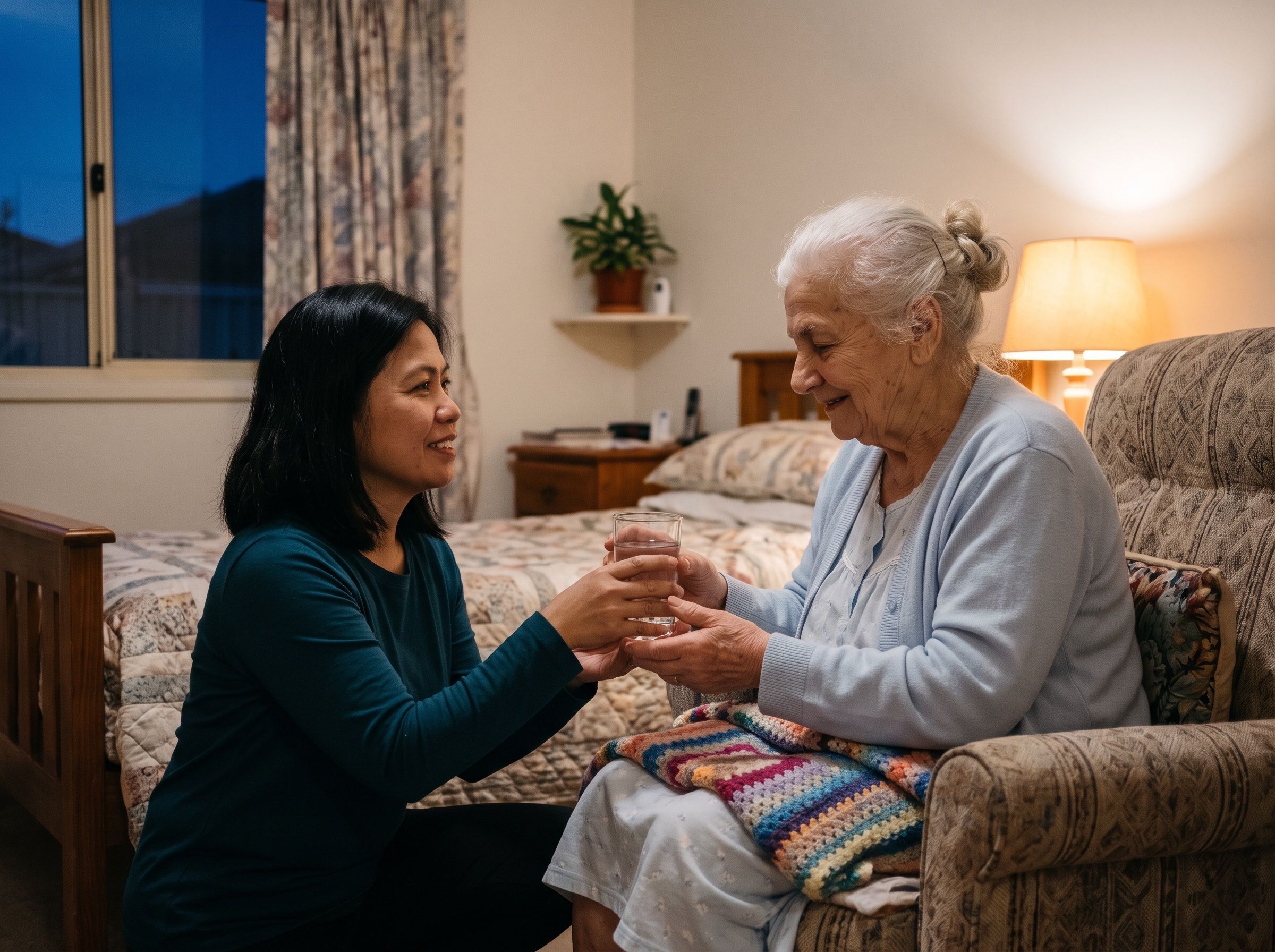 A support worker checks in with an older woman in her bedroom in the evening, by lamp light.
